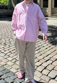 Světle růžová pruhovaná košile na knoflíky, volný střih, nošená přes bílé tričko; spárovaná s béžovými kalhotami a růžovými teniskami s bílými tkaničkami.