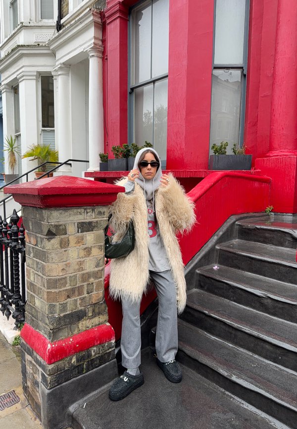 Osoba u sivoj dukserici s kapuljačom, bež krznenom kaputu, sivim hlačama, crnim cipelama i sunčanim naočalama stoji kraj stepenica crveno-niné kuće s biljkama na prozorskoj dasci.