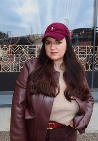 Weinrote Cordkappe, braune Lederjacke, beiger Pullover, roter Gürtel mit goldener Schnalle. Lange dunkle Haare, stehend vor gemustertem Glas.