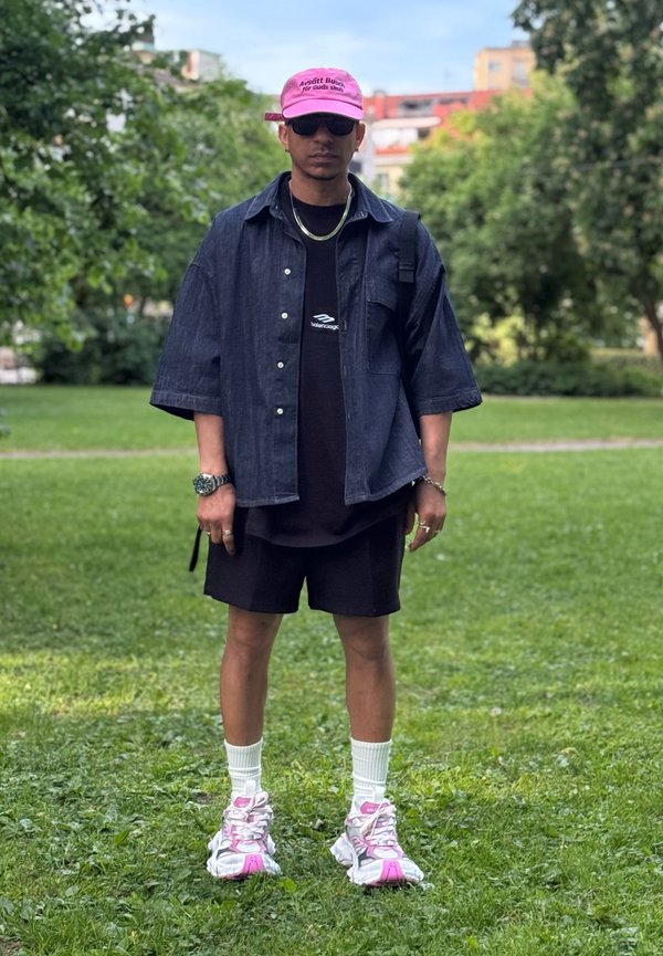Mann mit rosa Kappe, schwarzer Sonnenbrille, dunkelblauem Jeanshemd, schwarzen Shorts, weißen Socken und rosa-weißen Turnschuhen, der auf Gras in einem Park steht.