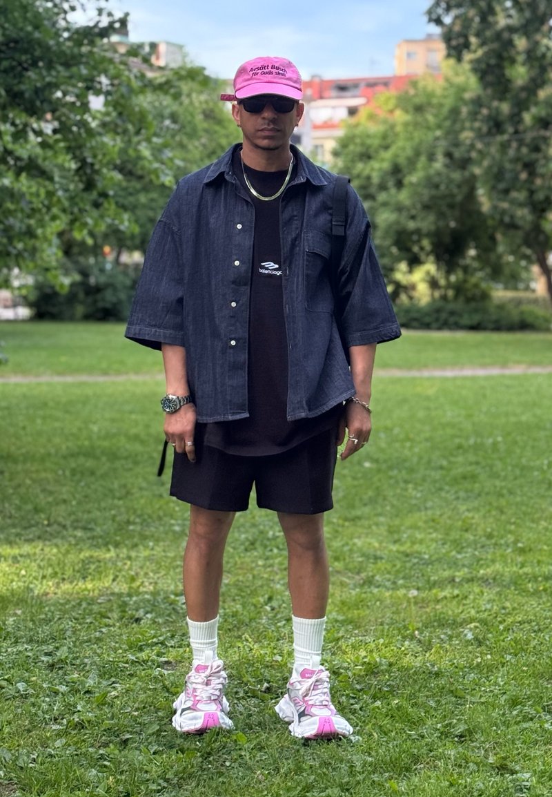 Homme portant une casquette rose, des lunettes de soleil noires, une chemise en jean foncée, un short noir, des chaussettes blanches et des baskets roses et blanches, debout sur l’herbe dans un parc.