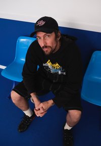 Sweatshirt preto com capuz e estampado, calções pretos e ténis pretos. Um boné escuro com logótipo rosa. Cadeiras de plástico azuis e fundo de chão azul.