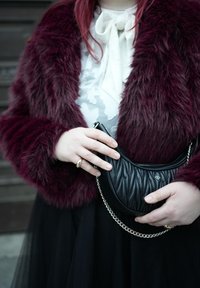 Άτομο που φοράει μπορντό γούνινο παλτό, λευκό πουκάμισο με φιόγκο, μαύρη φούστα, κρατώντας μια μαύρη καπιτονέ τσάντα χειρός με ασημένιο λουράκι αλυσίδας.