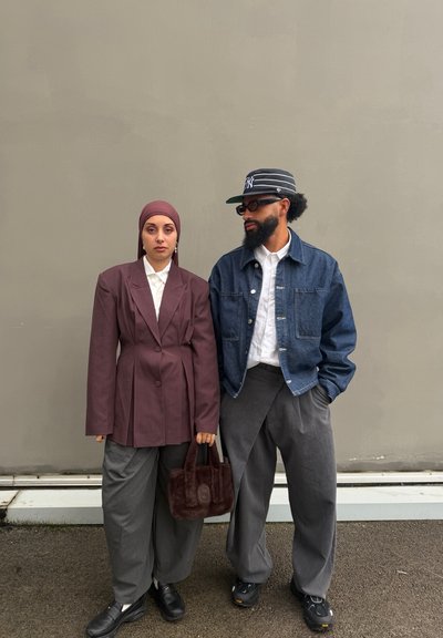 Dos personas están apoyadas contra una pared beige; una lleva un blazer granate y un pañuelo en la cabeza, sosteniendo una bolsa marrón, la otra lleva una chaqueta vaquera y una gorra con gafas.