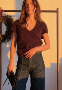 Femme portant un pull court à manches courtes en peluche de couleur violet foncé et un jean taille haute, tenant une pochette noire, debout près d'une couronne sous une lumière chaude.