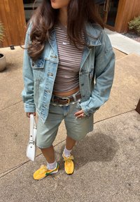 Denimjakke, stripete brun crop top, lyse denimshorts, brun belte med detaljer, og gule joggesko med grønn og sløyfedetaljer. Hvit håndveske.