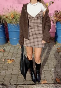 Smeđi teksturirani oversized kardigan preko bijele košulje, upareno sa smeđom kariranoj mini haljinom i crnim visokim čizmama s vrhovima. Crna torba za nošenje.