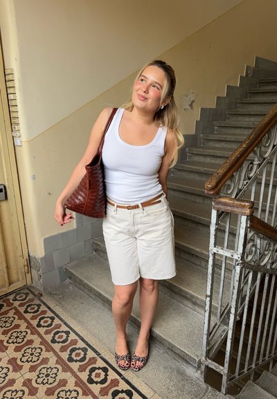 Top sin mangas blanco, pantalones cortos beige y zapatos planos con estampado de leopardo. Bolsa de mano trenzada marrón. Fondo de escalera con un suelo decorativo y con patrones.