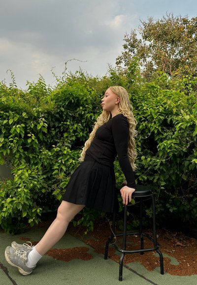 Top negro de manga larga con canalé y falda plisada negra, combinados con zapatillas grises. El fondo presenta un exuberante follaje verde y un taburete negro.