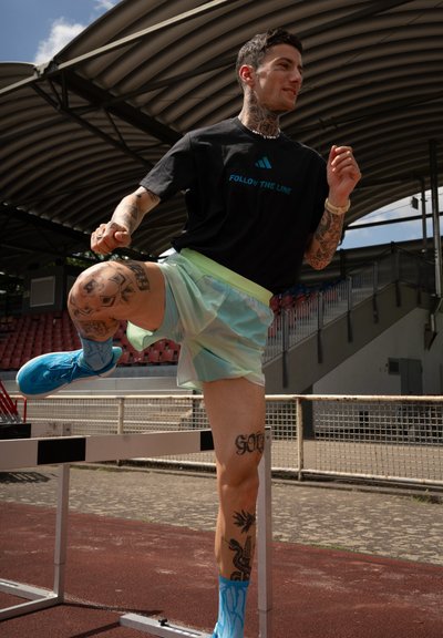 Ropa deportiva que presenta una camiseta negra con texto turquesa, pantalones cortos verde menta y zapatillas brillantes azules, acompañadas de tatuajes visibles.