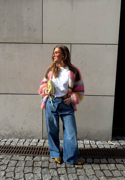 Mujer con un cárdigan de rayas de gran tamaño, camiseta blanca, vaqueros anchos azules, zapatos amarillos y sosteniendo un bolso amarillo, de pie contra una pared de hormigón.