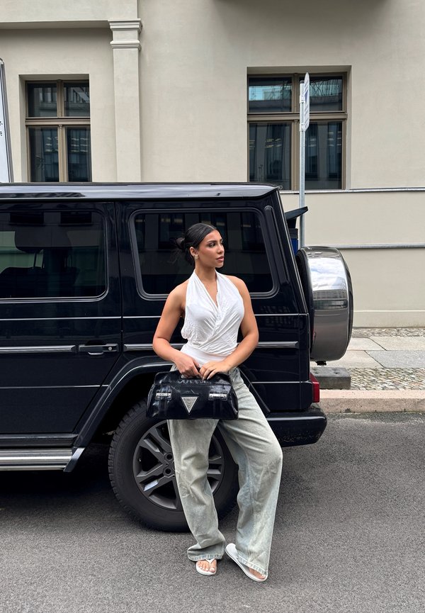 Bolsa preta da Prada com ferragens prateadas em uma modelo, vestindo um top halter branco e calças largas cinzas, ao lado de um SUV preto.
