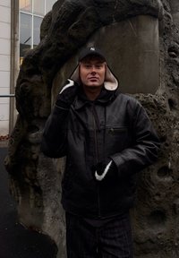 Jachetă de piele neagră cu fermoar, șapcă neagră și mănuși cu accente albe. Stând în fața unui fundal texturat din piatră gri.