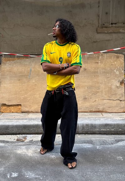 Hombre con cabello rizado que lleva una camiseta de fútbol de Brasil amarilla, pantalones anchos negros, chanclas, gafas de sol y pulseras, de pie con los brazos cruzados en la calle.
