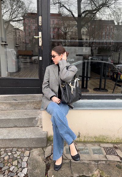 Blazer de tweed gris, suéter de cuello alto negro, jeans de corte ancho azules y bailarinas negras. Bolso negro con acentos blancos, sentado en unos escalones de piedra junto a un café.