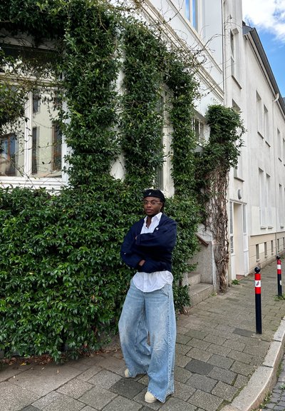 Persona vistiendo un suéter grande de color azul oscuro, camisa blanca y jeans anchos de color azul claro, de pie contra una pared cubierta de hiedra verde.
