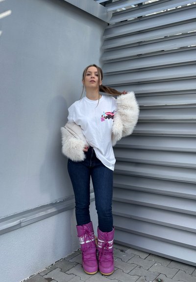 Camiseta blanca de gran tamaño con estampado floral, mangas de piel sintética clara, jeans azules y botas de nieve rosas con detalles blancos, de pie contra una pared gris.