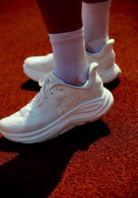 Balti sportiniai bateliai su tekstūruotu tinkliniu viršumi, suapvalintu padu ir HOKA ženklu; avimi su baltais kojinėmis ant rausvo paviršiaus.