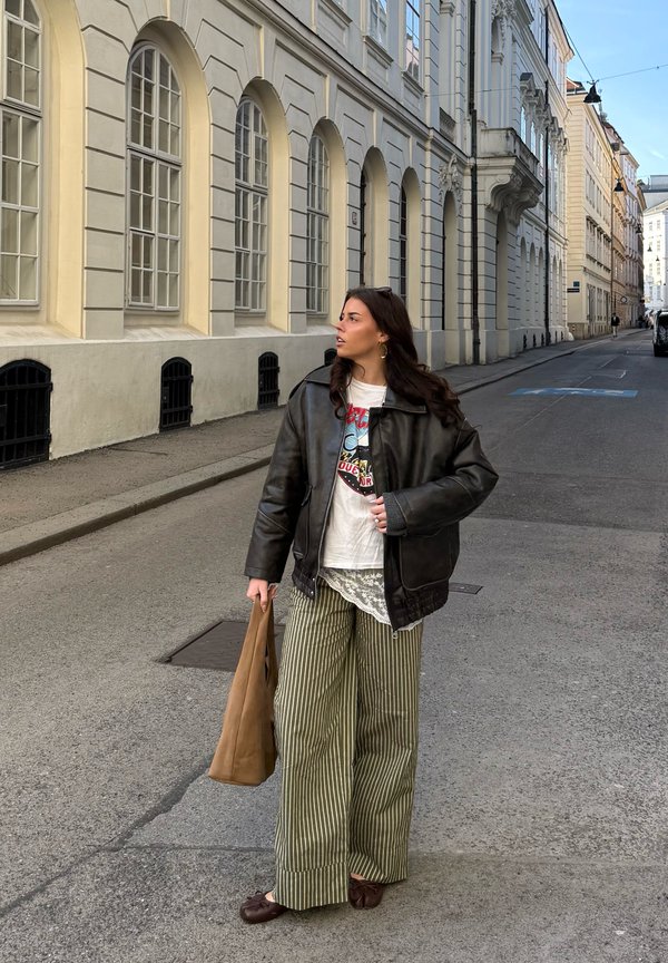 Kvinde i oversized sort læderjakke og stribede vide bukser, der holder en brun taske og står på en stille gade med høje historiske bygninger.