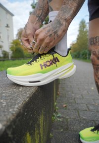 Osoba s tetovaniami zaväzuje šnúrky na jasne žltých a zelených bežeckých topánkach Hoka na kamennej hrane vonku.