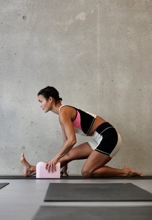 Esterilla de yoga con una superficie negra. Una persona vestida con un atuendo atlético en blanco y negro utiliza un bloque de yoga rosa mientras se estira hacia adelante. Pared de concreto liso.
