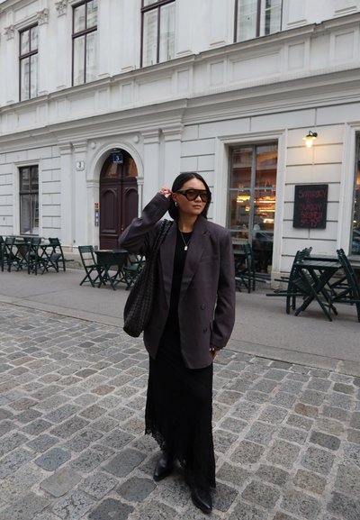 Blazer gris de gran tamaño sobre un vestido maxi negro con dobladillo de encaje, combinado con botines negros y gafas de sol oscuras. Un bolso de tela acentúa el conjunto.