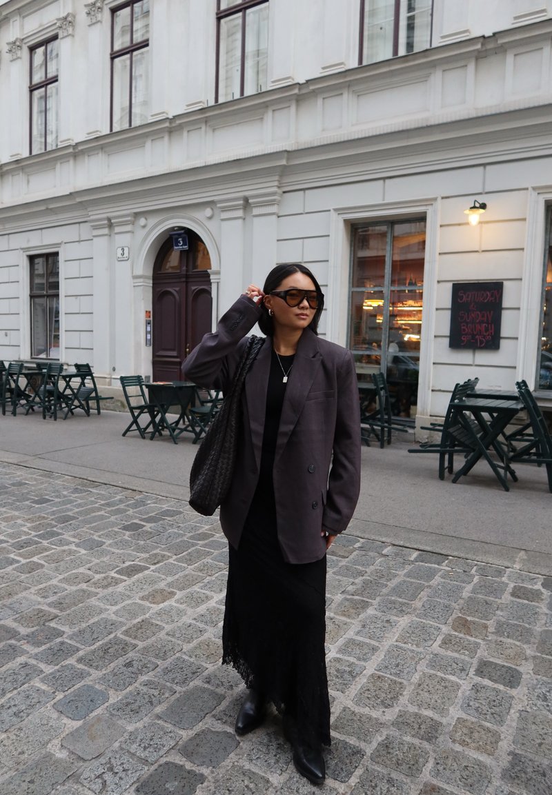 Oversized gray blazer over a black, lace-hemmed maxi dress, paired with black ankle boots and dark sunglasses. Woven handbag accentuates the look.