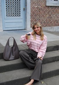 Rosa gestreiftes Langarmhemd, dunkle weite Hose, geflochtene braune Handtasche, hält eine Tasse Kaffee, sitzt auf grauen Steinstufen.