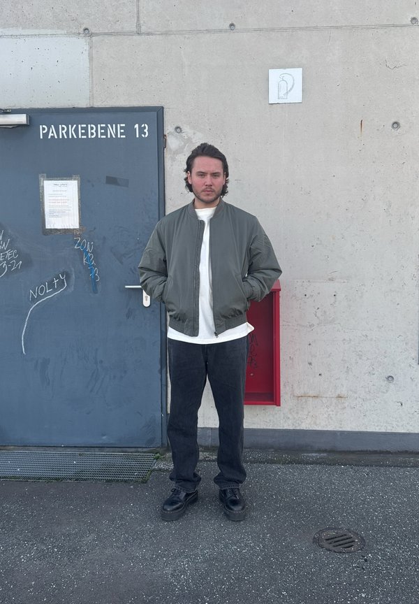 Olivovo-zelená bomber bunda, biele tričko, čierne džínsy, čierne topánky, postavený pred modrými dverami s bielym textom a červenou poštovou schránkou.