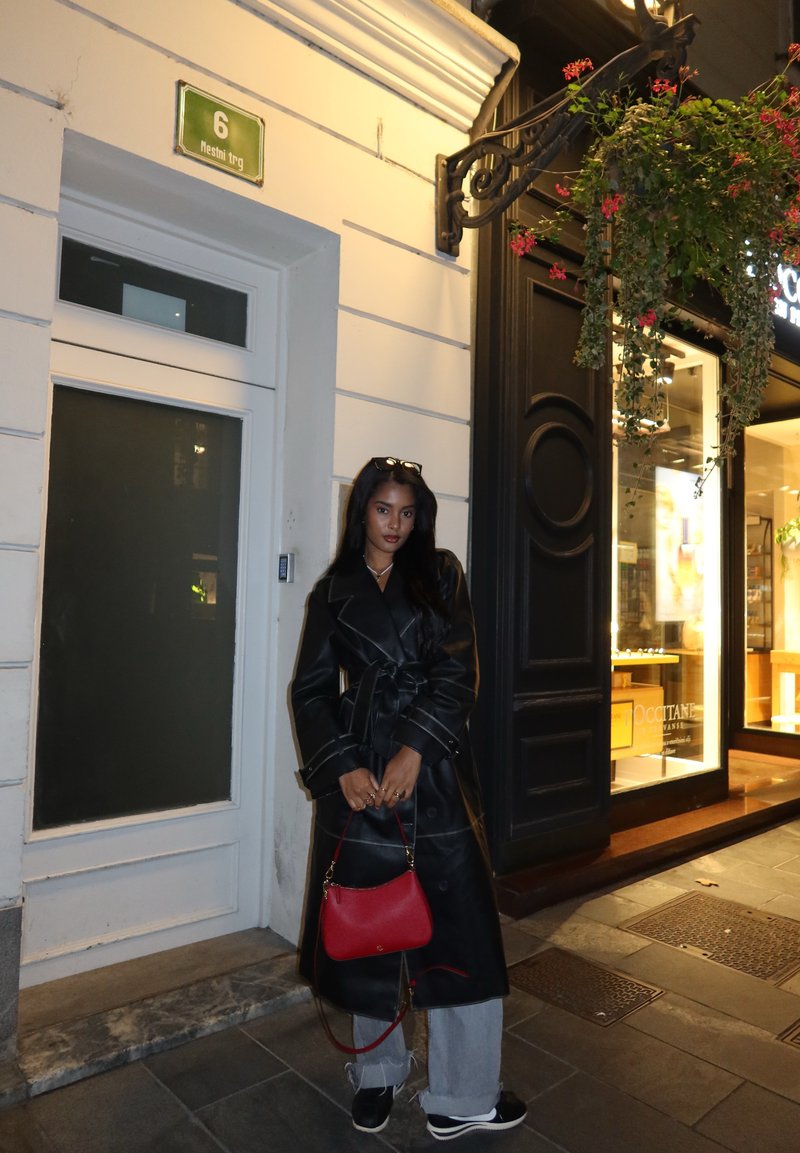 Μαύρο δερμάτινο παλτό με ζώνη στη μέση, γκρι παντελόνι φαρδύ, και κόκκινη τσάντα. Νυχτερινό τοπίο με κατάστημα και πράσινα φυτά στο φόντο.