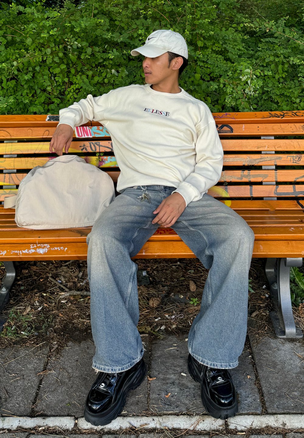 Beige Langarmhemd mit Logo, hellblaue baggy Jeans und schwarze Plateauschuhe. Im Hintergrund sind Grünanlagen und eine Holzbank zu sehen.