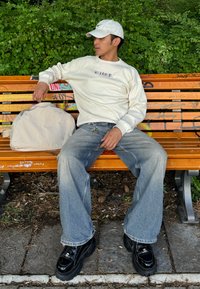 Beige Langarmhemd mit Logo, hellblaue baggy Jeans und schwarze Plateauschuhe. Im Hintergrund sind Grünanlagen und eine Holzbank zu sehen.