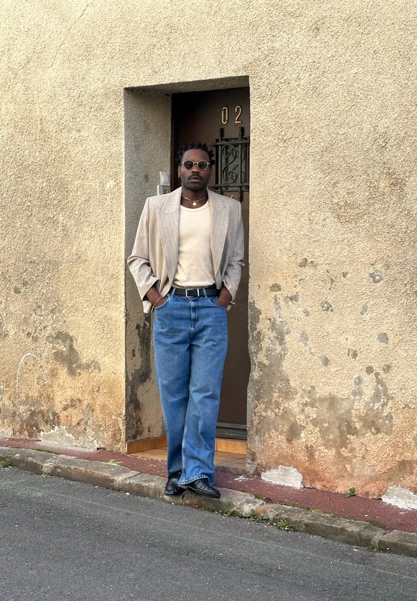 Mann med solbriller, beige blazer, kremfarget skjorte og blå jeans som lener seg mot en slitt vegg ved siden av en brun dør merket med nummer 02.