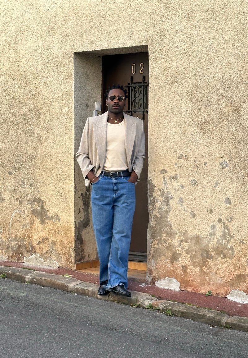 Άνδρας με γυαλιά ηλίου, μπεζ σακάκι, κρεμ πουκάμισο και μπλε τζιν, ακουμπώντας σε έναν φθαρμένο τοίχο δίπλα σε μια καφέ πόρτα με αριθμό 02.