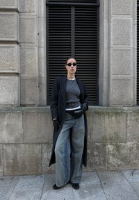 Femme portant un long manteau noir, un cardigan gris, un jean à jambes larges, des gants noirs, des lunettes de soleil noires et un sac banane noir, debout contre un mur de pierre.