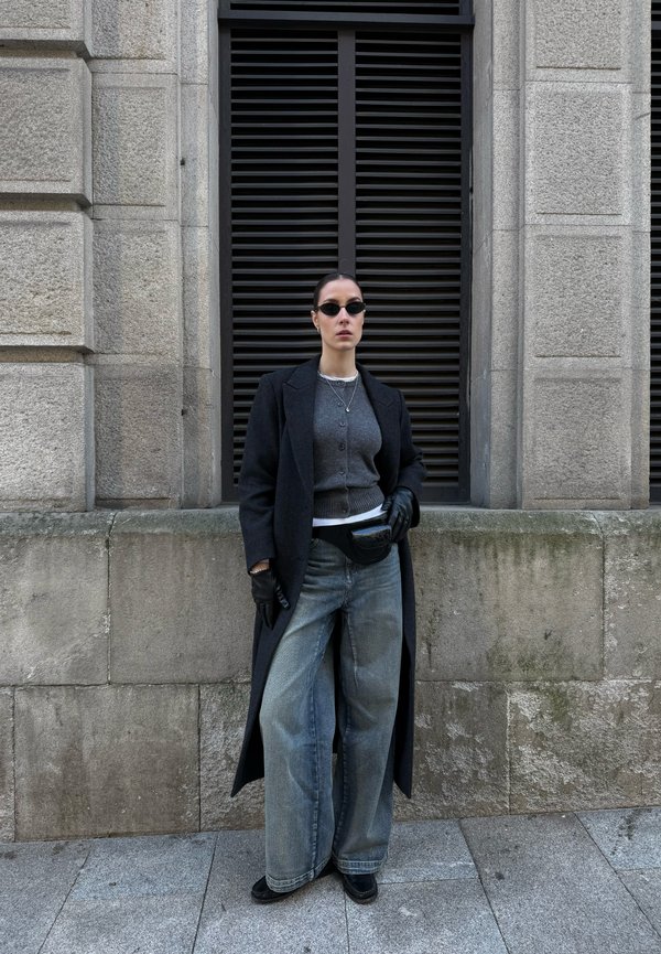 Ženska, oblečena v črni dolg plašč, siv pulover, široke kavbojke, črne rokavice, črna sončna očala in črna torbica okoli pasu, stoji ob kamnitem zidu.