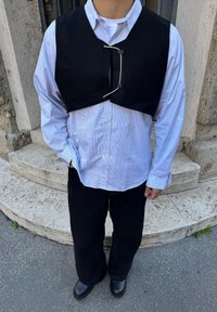 Personne portant un gilet court noir sur une chemise à rayures bleu clair à boutons, un pantalon noir et des chaussures noires, debout sur des marches en pierre.
