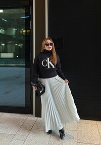 Pull noir côtelé avec logo "CK", jupe midi plissée blanche, bottines à talons noires et sac à main noir avec détail "M". Des lunettes de soleil complètent le look.