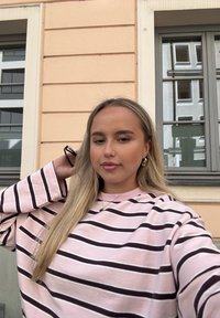 Oversized lyserød sweatshirt med mørkebrune striber, ribbet krave og lange ærmer. Modellen har langt hår og bærer en simpel halskæde.