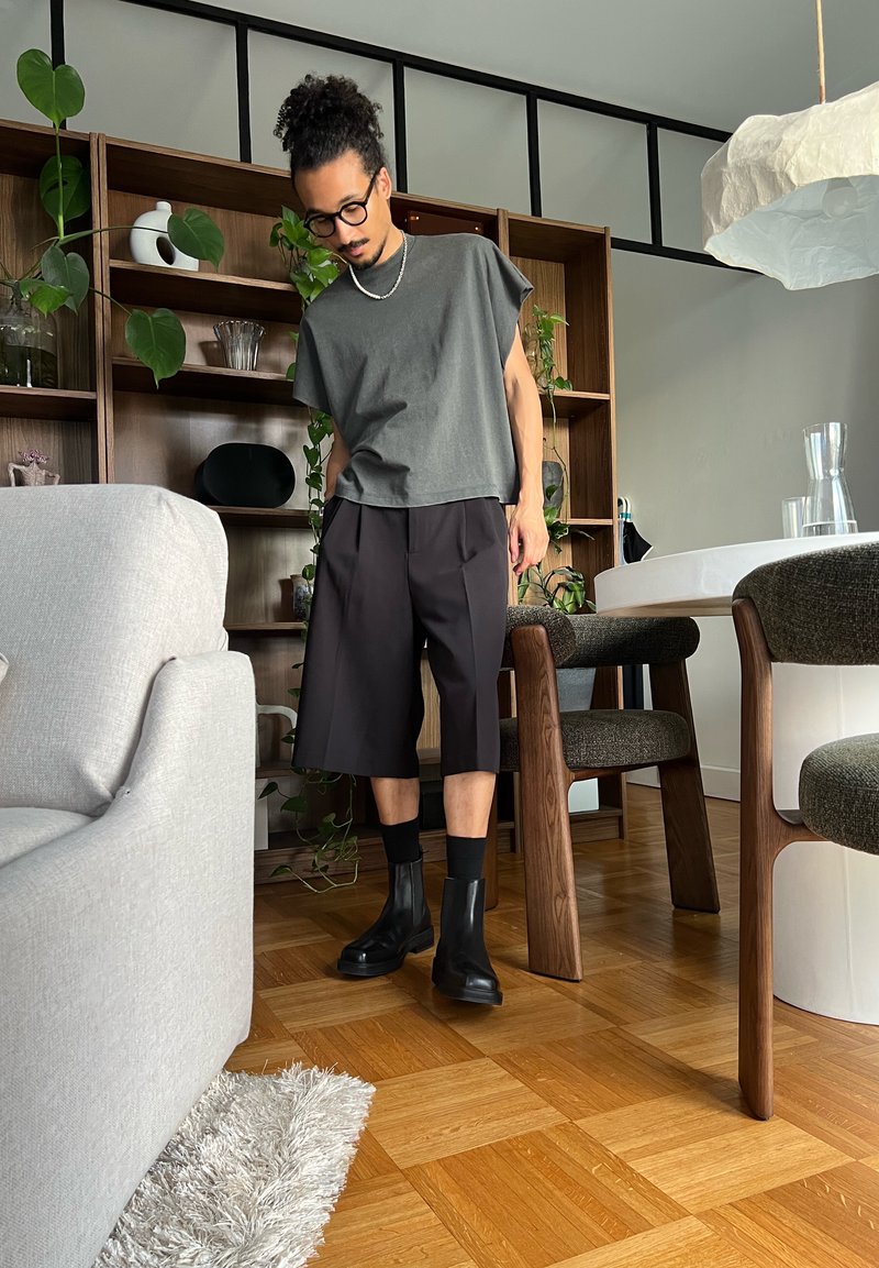 Homme portant des lunettes et les cheveux bouclés attachés, vêtu d'une chemise grise, d'un short noir et de bottes, se tient dans un salon moderne avec un sol en bois et des meubles.