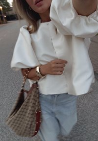 Biely blúz s naliatymi rukávmi a detailmi na gombíky, v kombinácii so vzorovanou hnedou kabelkou a svetlomodrými džínsami.