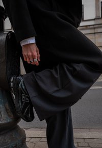 Baskets noires New Balance avec dessus texturé, associées à un pantalon gris et un long manteau noir. Main avec des bagues argentées reposant sur le genou.