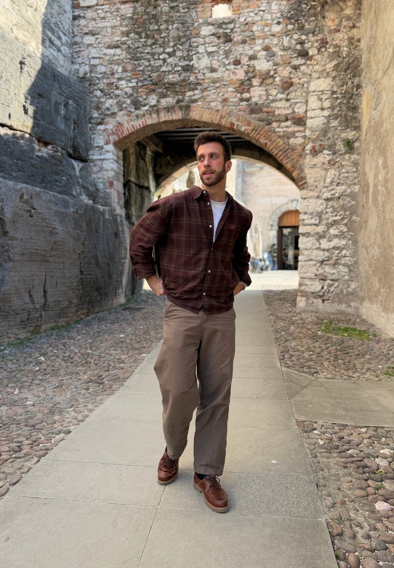 Man in brown plaid shirt and beige pants walking on stone path under old brick archway in historic setting.