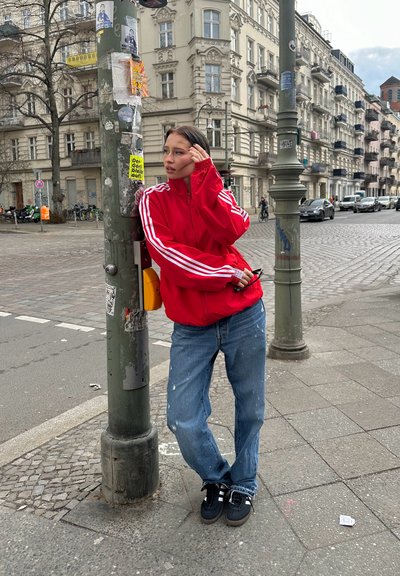 Chaqueta roja con cremallera y rayas blancas, jeans azules desgastados, zapatillas negras. De pie junto a una farola en un entorno urbano.