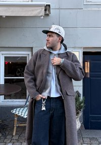 Abrigo largo marrón con estampado pata de gallo sobre una sudadera gris con cremallera, combinado con vaqueros oscuros. Lleva una gorra de béisbol y tiene llaves visibles en un llavero.