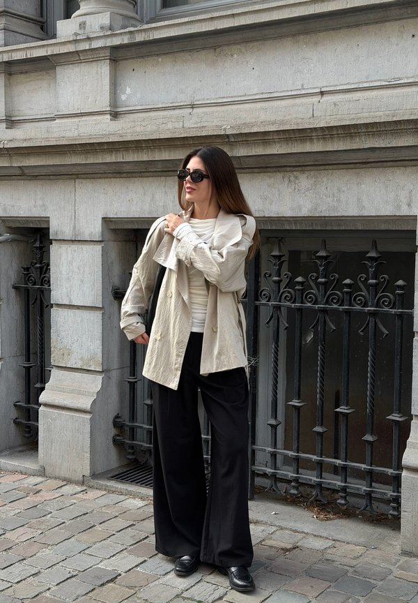 Mujer con gafas de sol, chaqueta beige, top blanco y pantalones anchos negros de pie en una calle adoquinada cerca de un edificio de piedra con barandilla de hierro forjado negra.