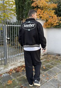 Must varsity jope valgete trükitähtedega seljal, soonikkoes mansettide ja alläärega, kombineeritud lahtiste mustade pükste ja tumedate tossudega.