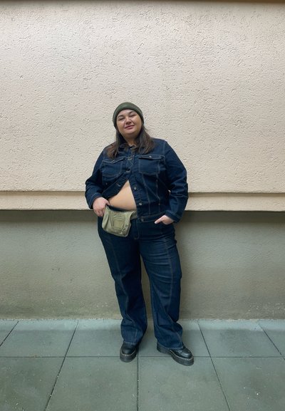 Chaqueta de denim y pantalones en azul oscuro, con un corte cropped. Gorro de lana verde oliva y una riñonera beige. Zapatos negros. Fondo de pared texturizada.