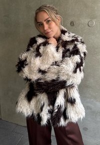 Fuskepelsjakke med en blanding av hvite og mørkebrune teksturer, med løs passform og beltedetalj, mot en nøytral bakgrunn.