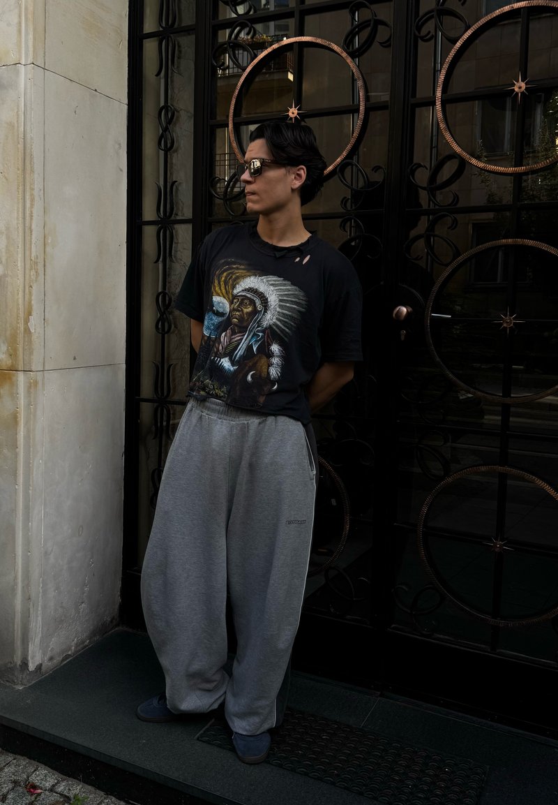 Giovane uomo che indossa occhiali da sole, pantaloni grigi oversize e una maglietta nera con grafica di un capo nativo americano, appoggiato a una porta metallica decorativa.